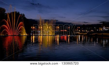 Amazing fantastic colorful fountain with bright illumination on the water pond or river with beautiful reflection at the evening or night. Beauty urban recreational concept.
