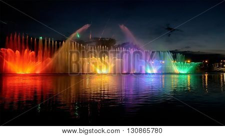Amazing fantastic colorful fountain with bright illumination on the water pond or river with beautiful reflection at the evening or night. Beauty urban recreational concept.