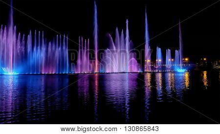 Amazing fantastic colorful fountain with bright illumination on the water pond or river with beautiful reflection at the evening or night. Beauty urban recreational concept.