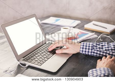 Man typing on laptop with blank white screen on wooden desktop with business report and coffee cup. Mock up