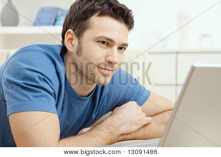 Felice giovane uomo posa sul divano e utilizzando il computer portatile a casa, sorridendo.