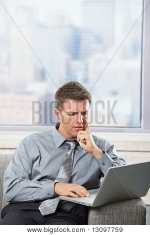 Biznesmen koncentruje się pracy na laptopa siedząc na kanapie w budynku scyscraper.