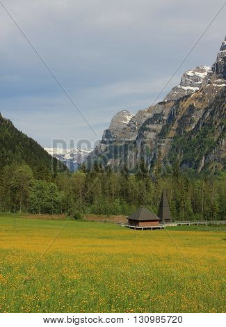 Meadow full of yellow wildflowers. High mountains of the Glaernisch. Modern church. Spring scene in the Swiss Alps.