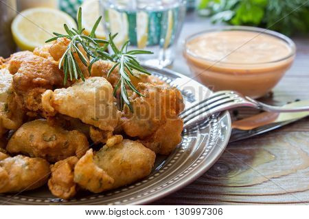 In the foreground shrimps in crispy dough on a plate with rosemary, cutlery, in the background glass of water, lemon, herbs, bowl with sauce. Shrimps in pastry for lunch. Horizontal. Close.