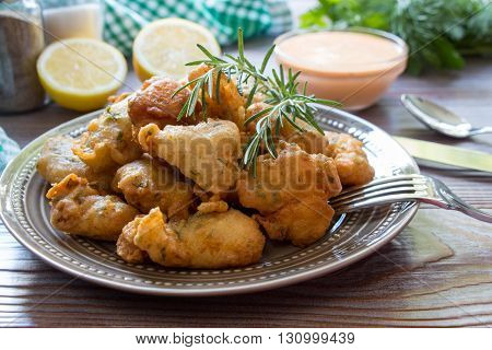 In the foreground shrimps in crispy dough on a plate with rosemary, cutlery, in the background lemon slices, salt, pepper, herbs, bowl with sauce. Shrimps in pastry for lunch. Horizontal. Close.