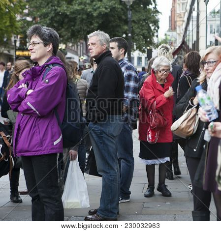 London, England - April 12, 2016 An Elderly Woman In A Red Cloak Is Making Her Way Through The Crowd