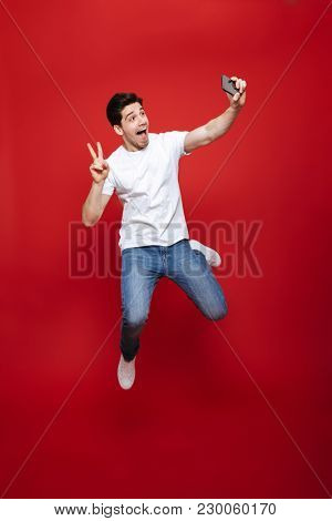Full length portrait of a cheerful young man in white t-shirt showing peace gesture while taking a selfie and jumping isolated over red background