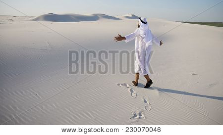Cheerful Muslim Guy Arabian Uae Sheikh Walks Along White Sand Of Wide Desert On Warm Summer Evening.