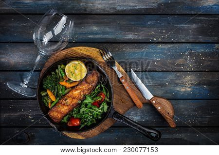 Grilled Salmon Fillet In A Frying Pan, Vegetables, Arugula With A Lemon Served With Cutlery And Glas