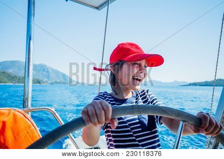 Cute Boy Captain On Board Of Sailing Yacht On Summer Cruise. Travel Adventure, Yachting With Child O