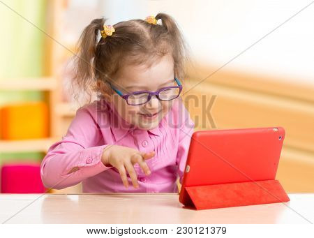Smart kid in spectacles using tablet PC or e-book sitting at table in her room