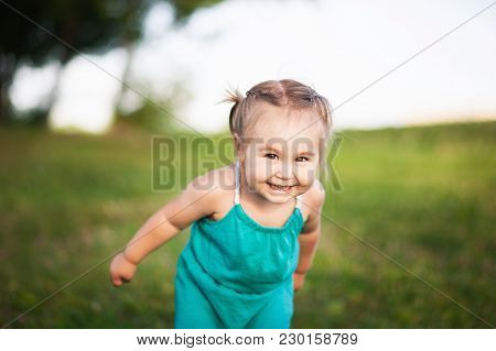 A Little Girl In A Green Sarafan Smiles In Summer Against A Background Of Green Young Grass. Unplugg