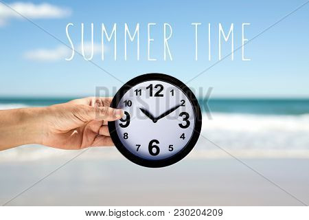 closeup of a young caucasian man with a clock in his hand on the beach, next to the seashore, and the text summer time