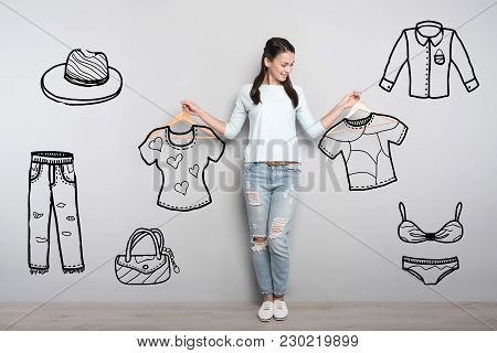 New Clothes. Beautiful Cheerful Student Feeling Interested While Standing With Wooden Hangers In Her