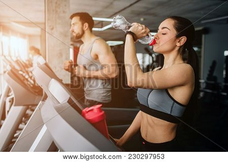 Beautiful And Well-built Girl Is Running On The Running Machine Besides Her Sport Partner And Drinki