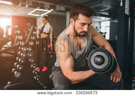 Close Up Of Strong Guy That Exercising With A Dumbbell On The Sport Bench While Hir Girlfriend Is St
