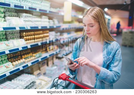 Beautiful And Attractive Girl Is Standing Near Shelf With Eggs In The Grossary Store. She Is Watchin