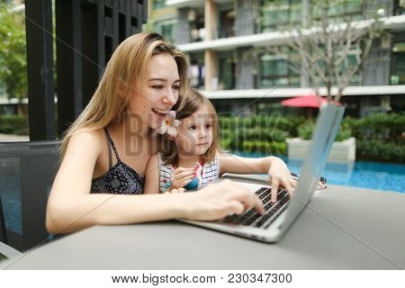 Happy Parent Mother And Daughter Use Laptop On Background Of Swimming Pool On Sunny Day. Concept Of 