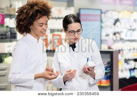 Two experienced female pharmacists wearing lab coats while analyzing together the package of a new pharmaceutical drug in the interior of a modern pharmacy