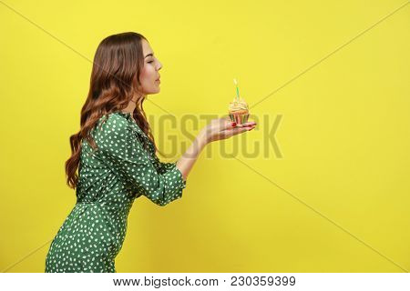 Beautiful young woman snuffing out candle on birthday cupcake against color background