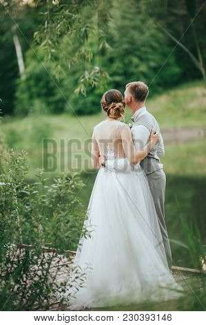 The Bride And Groom Walk On A Background Of Lake. Romantic Wedding Photo.