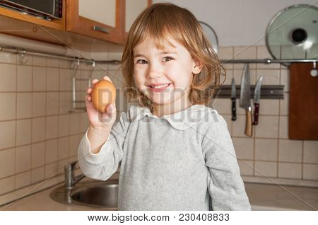 Little Baby Girl Holds Egg And Smiles On Kitchen Background