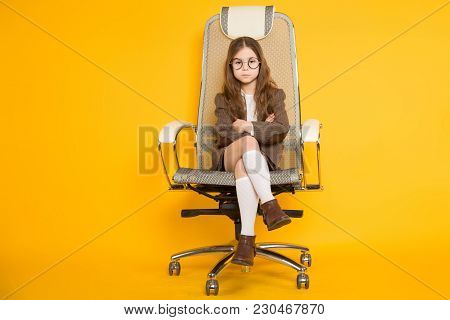 Portrait Of Long-haired Little Girl In Eyeglasses Sits In Armchair Wearing Brown Jacket, Shorts And 