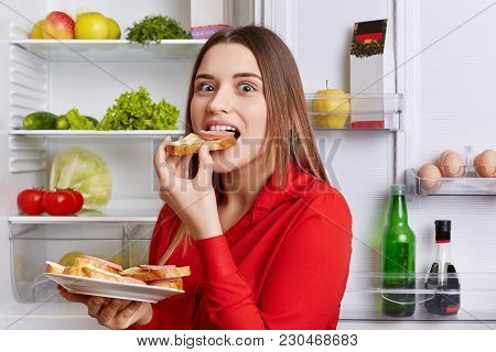 Hungry Adorable Woman Eats Delicious Sandwhiches With Cheese And Sausage, Stands Near Opened Refrige