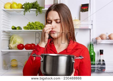 Young Brunette Woman With Displeased Expression Smells Spoiled Soup In Stew Pan, Feels Musty Smell A