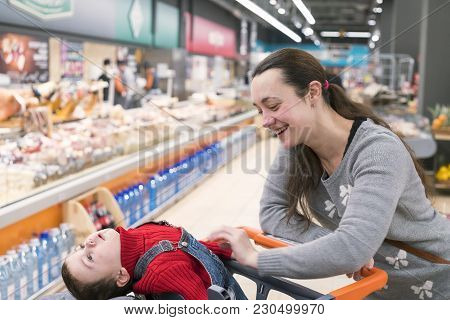 A Young Woman With A Small Son In A Supermarket. The Boy Sits In A Baby Cart And Laughs.