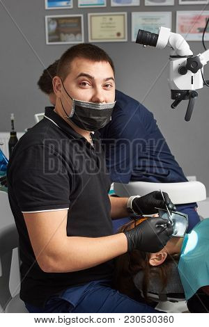 Young Dentist In A Black Mask And Gloves Treats A Patient Using A Cofferdam And A Microscope. Modern