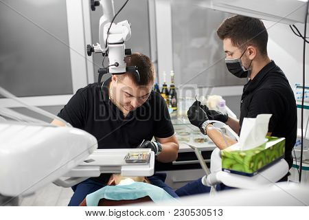 Two Male Dentists In Black Uniforms Are Working With Dental Instruments While Curing Teeth Of A Fema