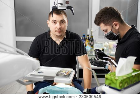 Male Dentist In A Black Uniform Is Looking To The Camera While Another Is Working With Dental Materi