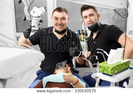 Two Male Dentists In Black Uniform Are Showing Thumbs Up And Smiling To The Camera While Sitting In