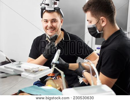 Male Dentist In A Black Uniform And Mask Performs Some Treatment For A Patient While Another One Smi
