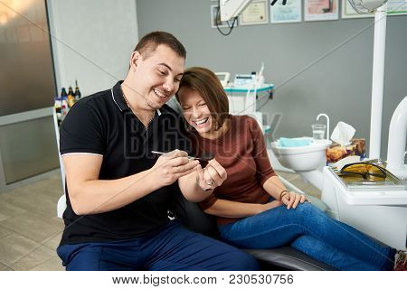 Male Dentist In A Black Uniform And His Patient Are Smiling While Looking At An X-ray Shot