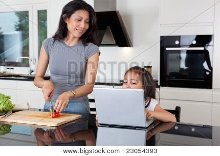 Jolie fille à l'aide d'ordinateur portable tandis que la mère coupe les légumes en cuisine