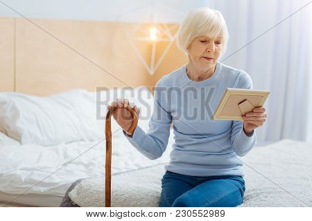 Aged Woman. Calm Serious Tired Aged Woman With A Wooden Walking Stick Sitting On Big Soft Bed And Th