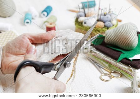 Closeup Hands Of The Needlewoman Hold Scissors And Cut Out A Piece Of Pattern For A Handmade Toy.