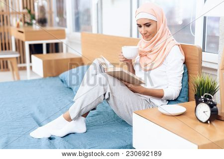 An Arabian Woman Is Drinking Tea And Reading A Book While Sitting On The Bed. She Rests At Home.