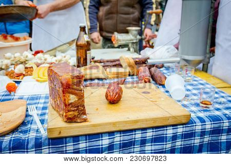 Headcheese And Smoked Sausage On A Wooden Chopping Board At Outdoor Kitchen, People In Background Ar