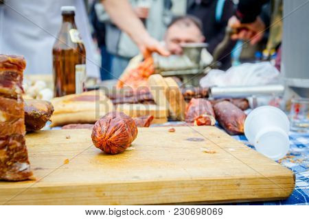 Headcheese And Smoked Sausage On A Wooden Chopping Board At Outdoor Kitchen, People In Background Ar