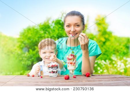 Mother And Her Son Child Boy Are Eating Laered Dessert In Sunny Day In Green Garden.