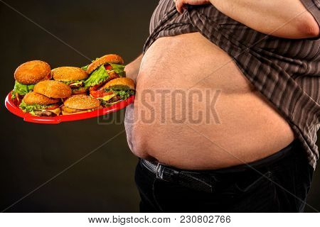 Man belly fat with hamberger group fast food. Breakfast for overweight person. Cropped shot of junk meal leads to obesity. Male lifted shirt, shows bare stomach. Health problems due to malnutrition.