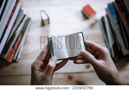 A Man Reading A Miniature Book Among The Big Books In The Library At The Wooden Table.
