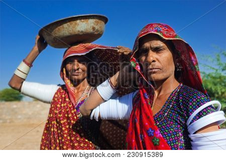 GODWAR REGION, INDIA - 14 FEBRUARY 2015: Rabari tribeswomen stand in field wearing sarees and upper-arm bracelets. One balances bucket on head. Rabari are an Indian community in the state of Gujarat.