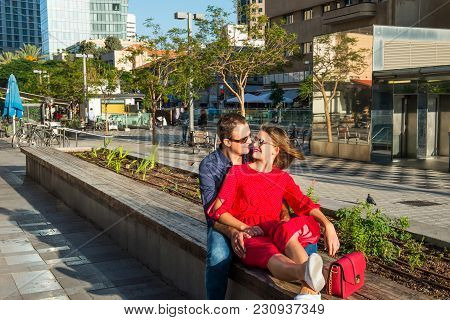 Young Loving Couple In Bright Clothes And Sunglasse Ssitting On The Bench In The City Area. Embracin