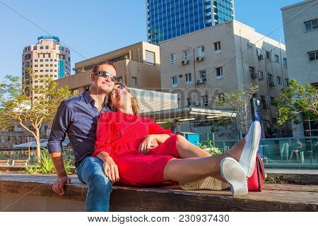 Young Loving Couple In Bright Clothes And Sunglasse Ssitting On The Bench In The City Area. Embracin