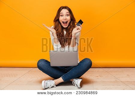 Surprised happy brunette woman in sweater sitting on the floor with laptop computer while holding credit card and looking at the camera over yellow background