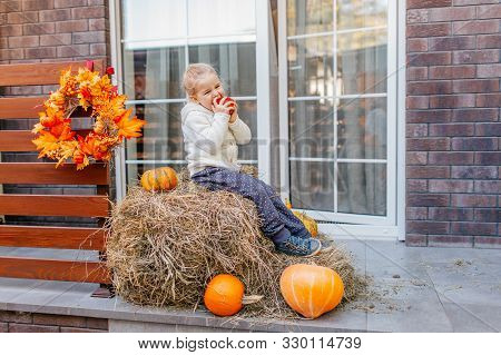 Adorable Smiling Caucasian Baby Toddler In White Knittes Jacket Sitting On The Haystack With Pumpkin
